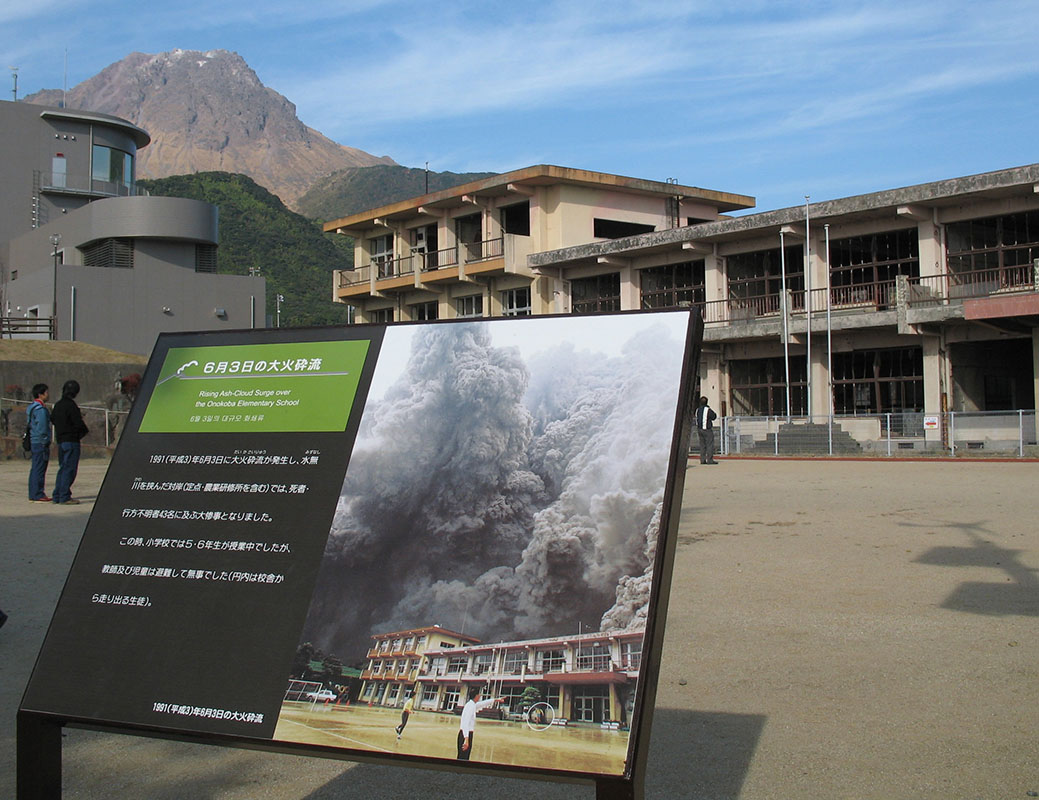 An image from the 1991 disaster at Mount Unzen, Japan, appears in signage near the elementary school. It was among the buildings overrun by the pyroclastic flow of June 3. In December 2007, a new armored observatory stood at left. Behind it, vents at the dome were still producing small steam emissions. (Kevin Scott photo)